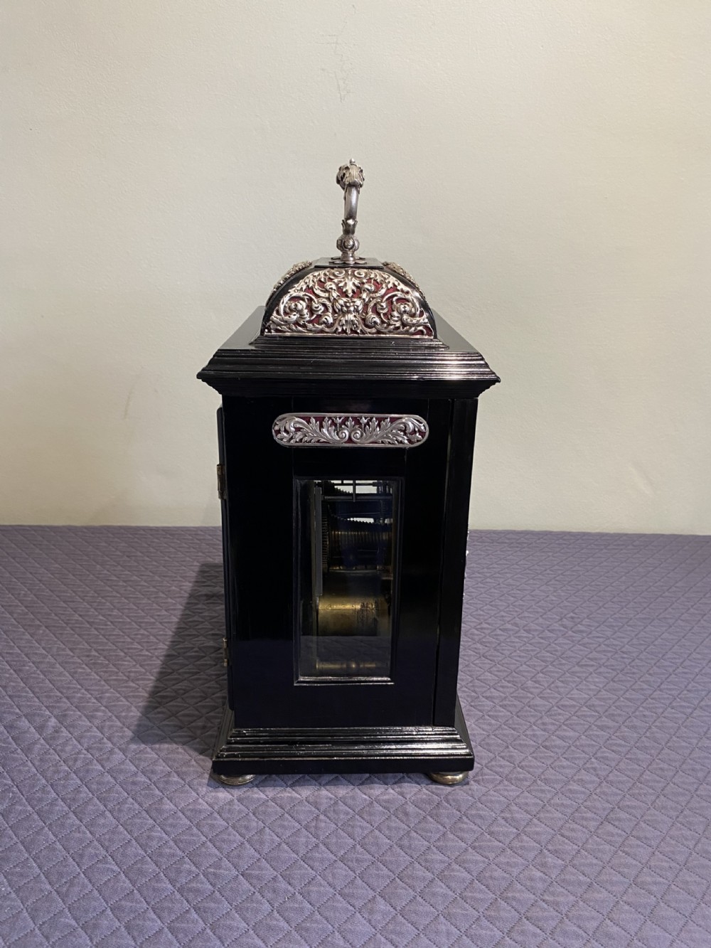 A late 17th-century black ebony table clock featuring repousse silver mounts, an ornate carrying handle, and a brass dial with silvered chapter ring. View VI