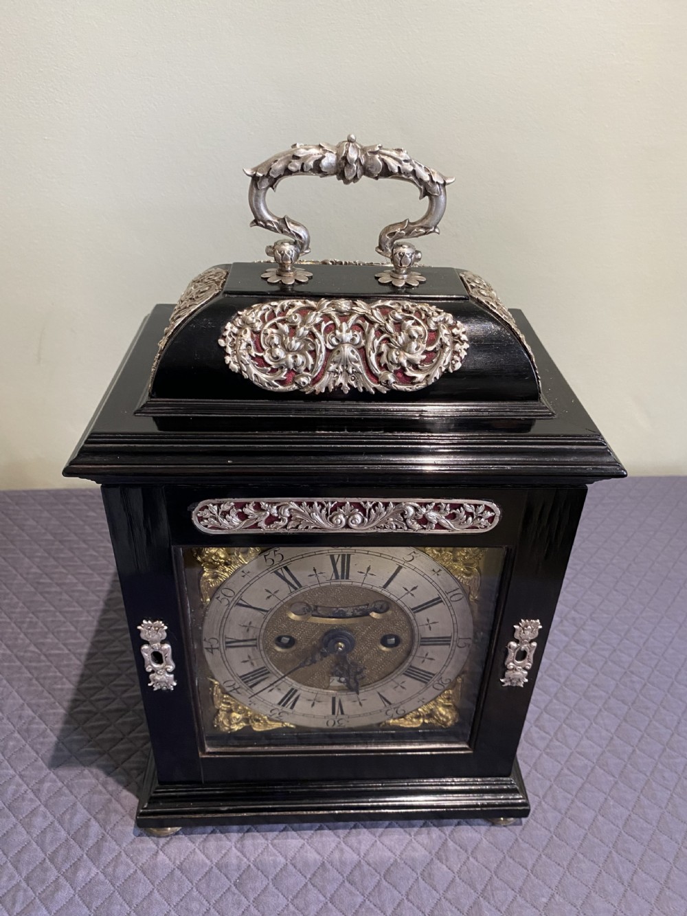 A late 17th-century black ebony table clock featuring repousse silver mounts, an ornate carrying handle, and a brass dial with silvered chapter ring. View VII