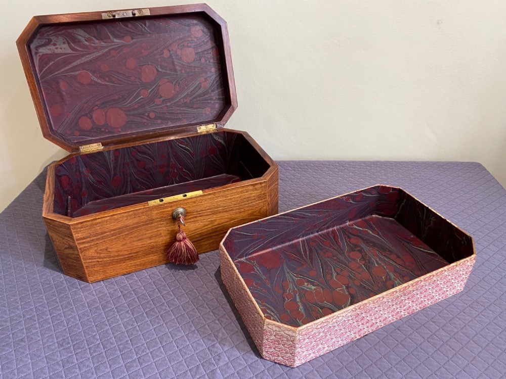 An open octagonal rosewood box featuring a removable tray, marbled paper lining in red and grey tones, and a key with a silk tassel.