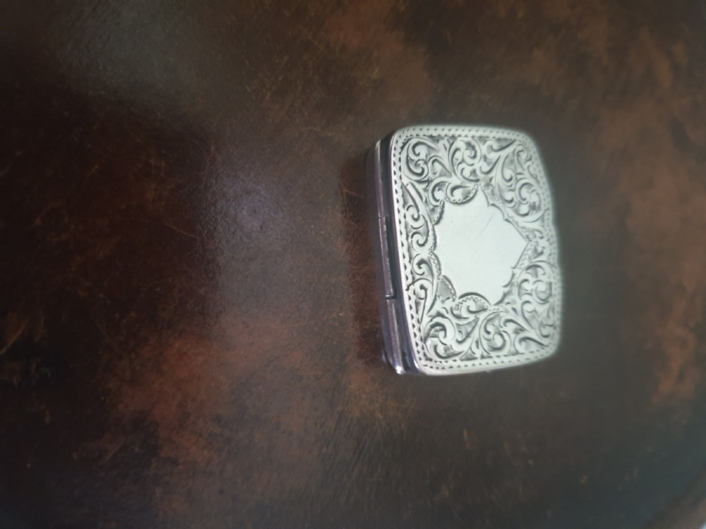 An Edwardian sterling silver rectangular pillbox featuring engraved foliate scrollwork and a central unengraved cartouche.