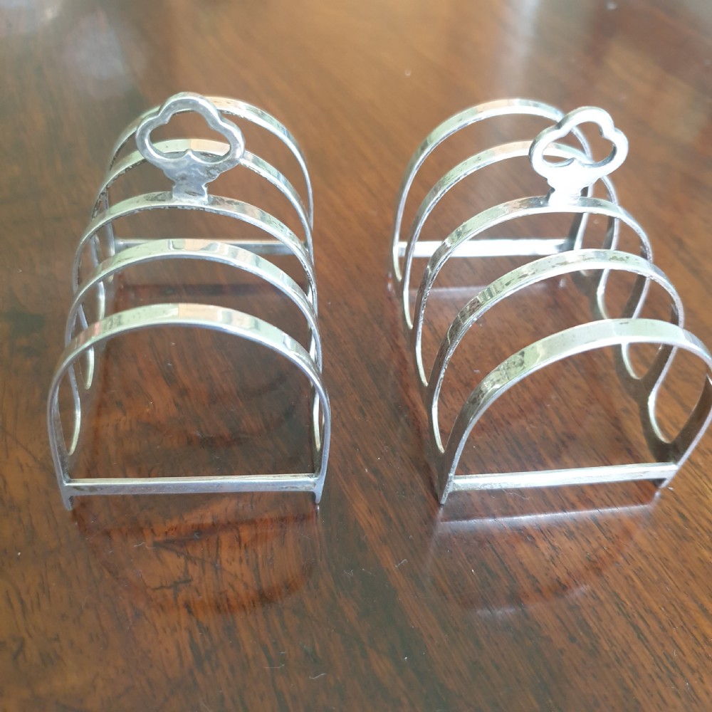 A pair of four-slot sterling silver toast racks featuring curved arches and trefoil central handles resting on a polished wooden surface.