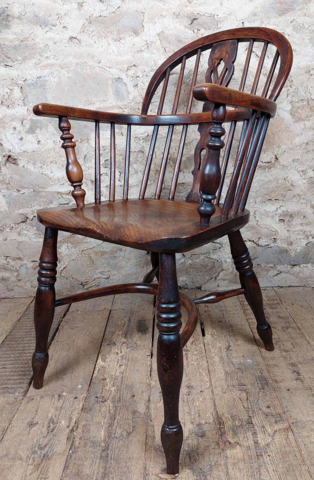 A dark brown wooden low back Windsor armchair featuring a shaped saddle seat, turned spindles, armrests, a pierced central back splat, and turned legs.