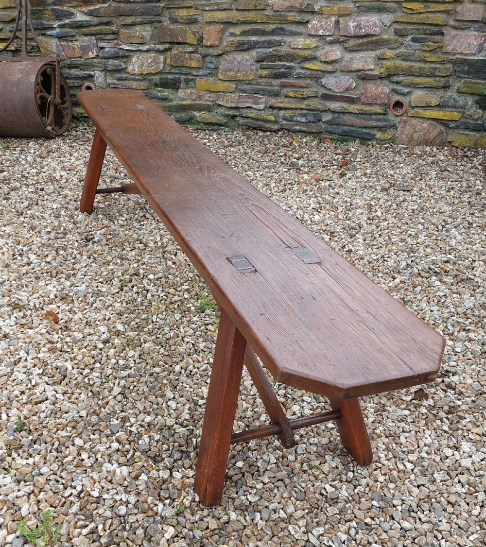 A long rustic pine bench with a flat timber top, splayed legs, and cross-braced support structure. View III