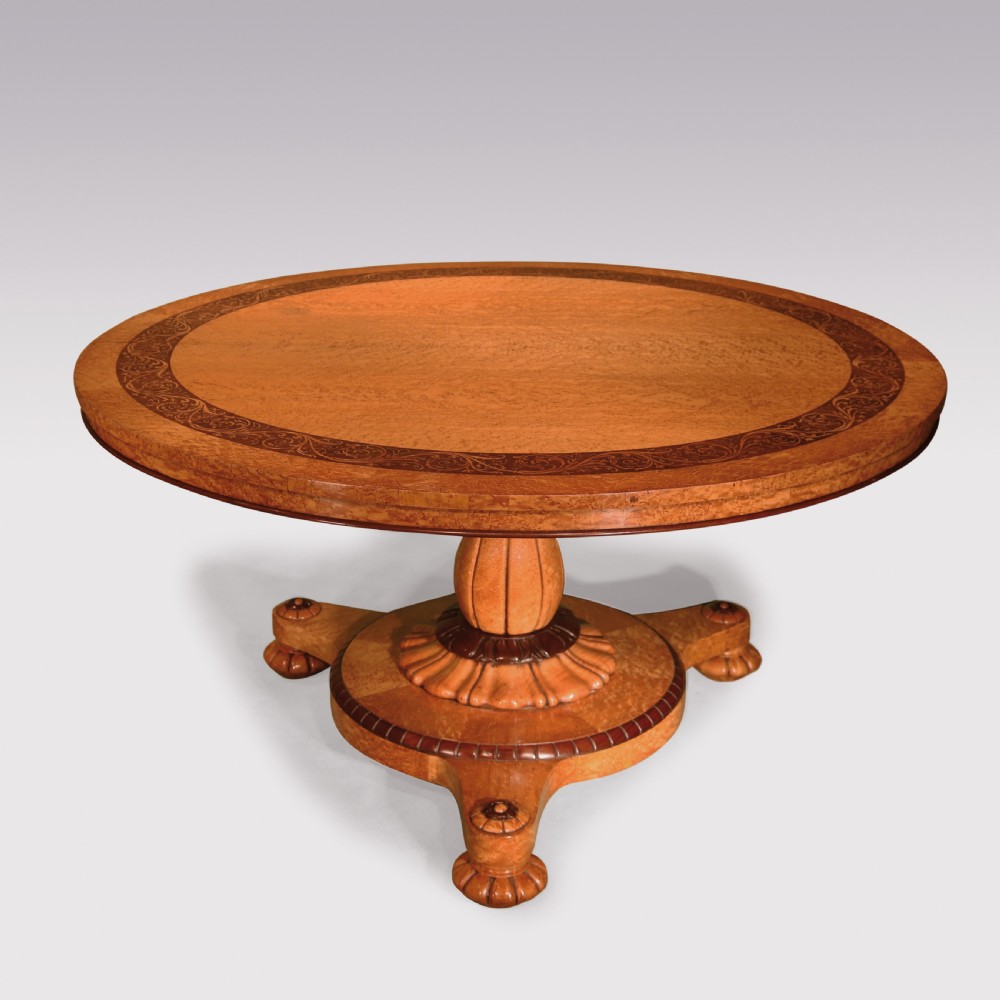 A circular mid-19th-century centre table crafted from birdseye maple featuring an inlaid decorative border, a carved baluster pedestal, and a platform base on carved feet.