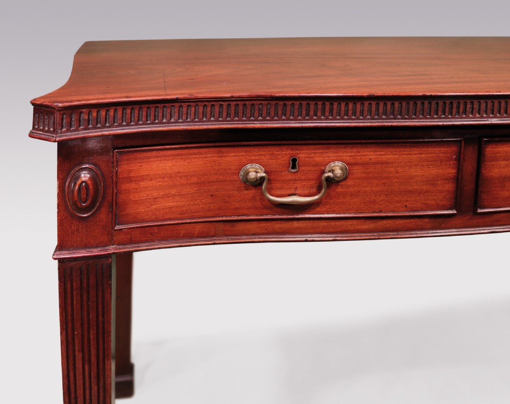 An 18th-century George III mahogany serving table featuring a serpentine front, three drawers with brass swan-neck handles, and fluted square tapered legs. View IV