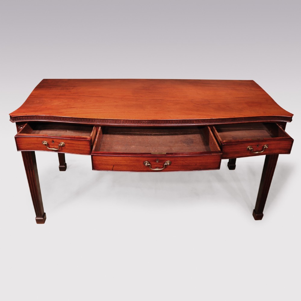 An 18th-century George III mahogany serving table featuring a serpentine front, three drawers with brass swan-neck handles, and fluted square tapered legs. View V