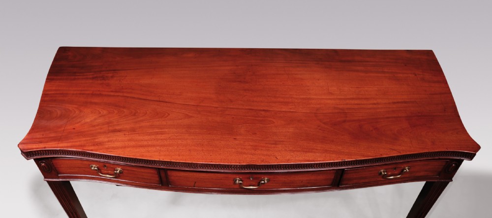 An 18th-century George III mahogany serving table featuring a serpentine front, three drawers with brass swan-neck handles, and fluted square tapered legs. View VI