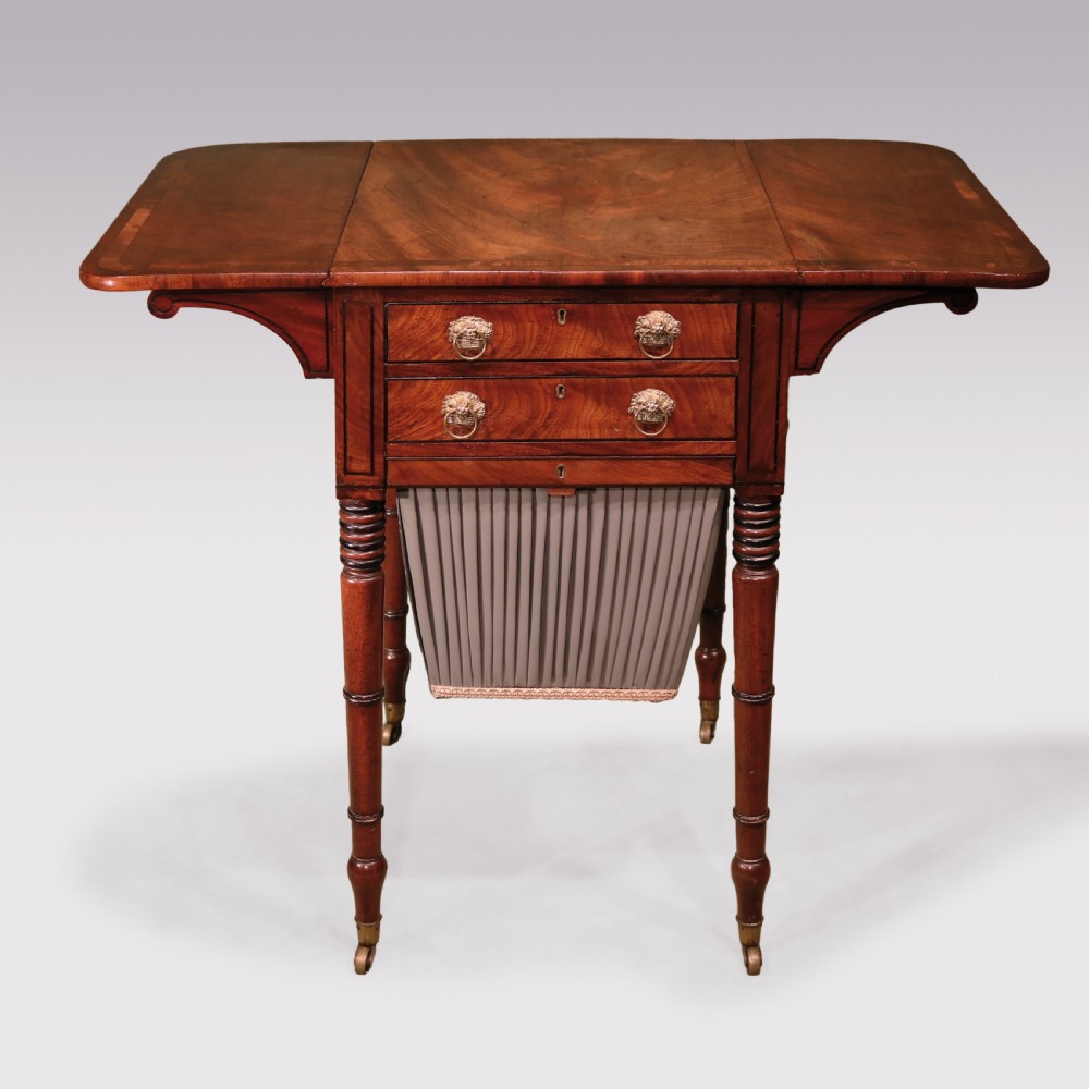A Regency mahogany work table featuring two drop-leaf sides, two front drawers with brass lion-head ring handles, a pleated grey fabric basket below, and reeded legs on brass castors.