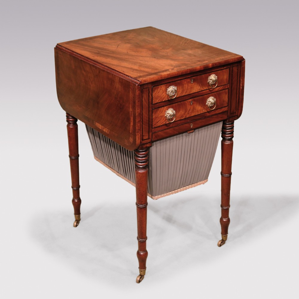 A Regency mahogany work table featuring two drop-leaf sides, two front drawers with brass lion-head ring handles, a pleated grey fabric basket below, and reeded legs on brass castors. View II