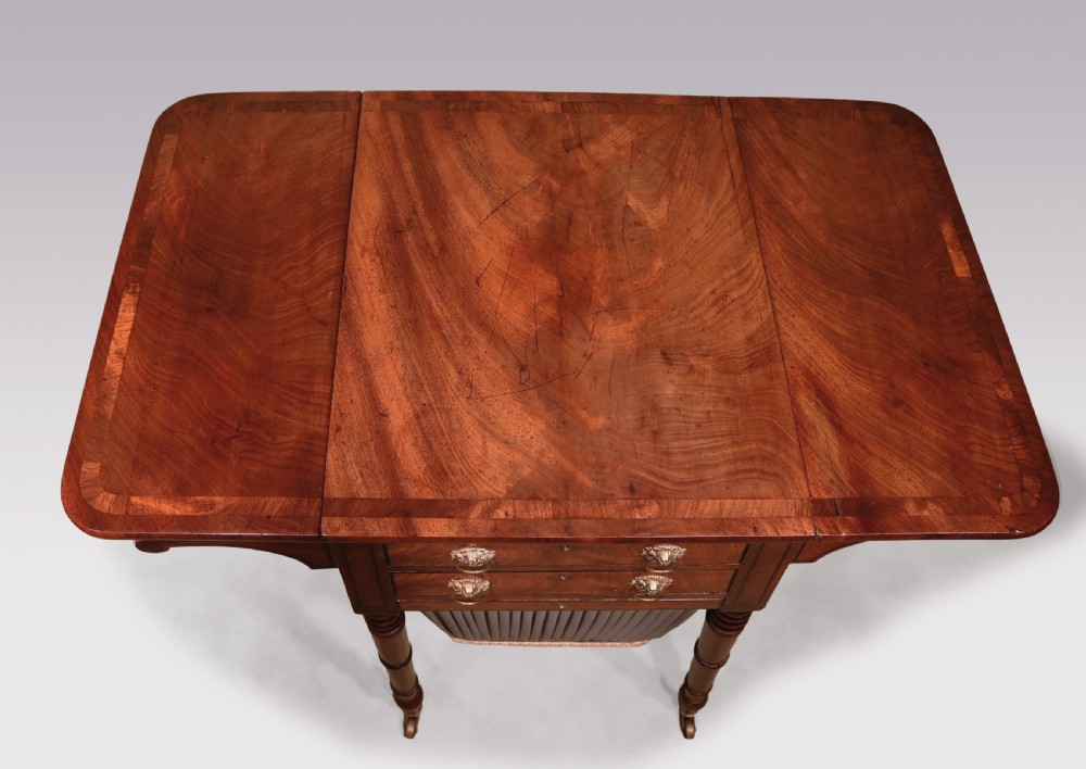 A Regency mahogany work table featuring two drop-leaf sides, two front drawers with brass lion-head ring handles, a pleated grey fabric basket below, and reeded legs on brass castors. View III