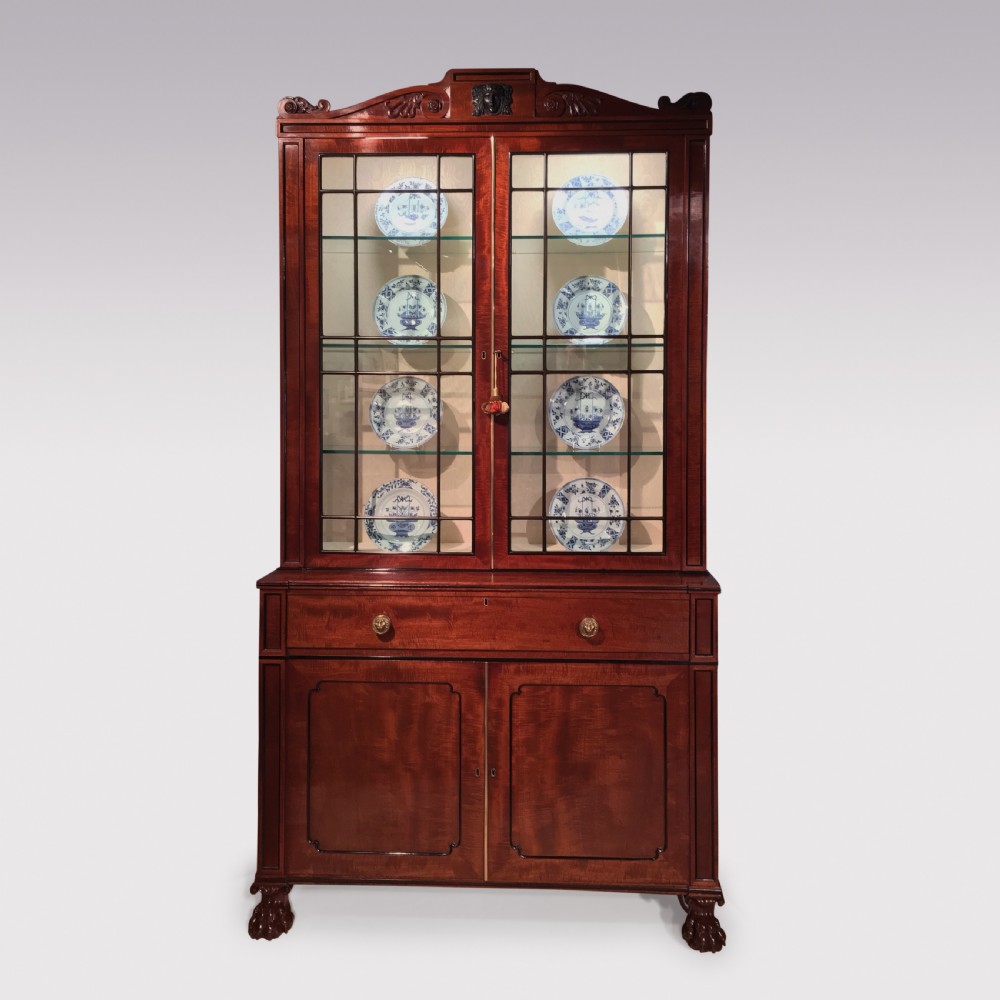 A Regency period mahogany secretaire bookcase featuring glazed upper doors displaying ceramics, a carved mask pediment, and carved lion paw feet.