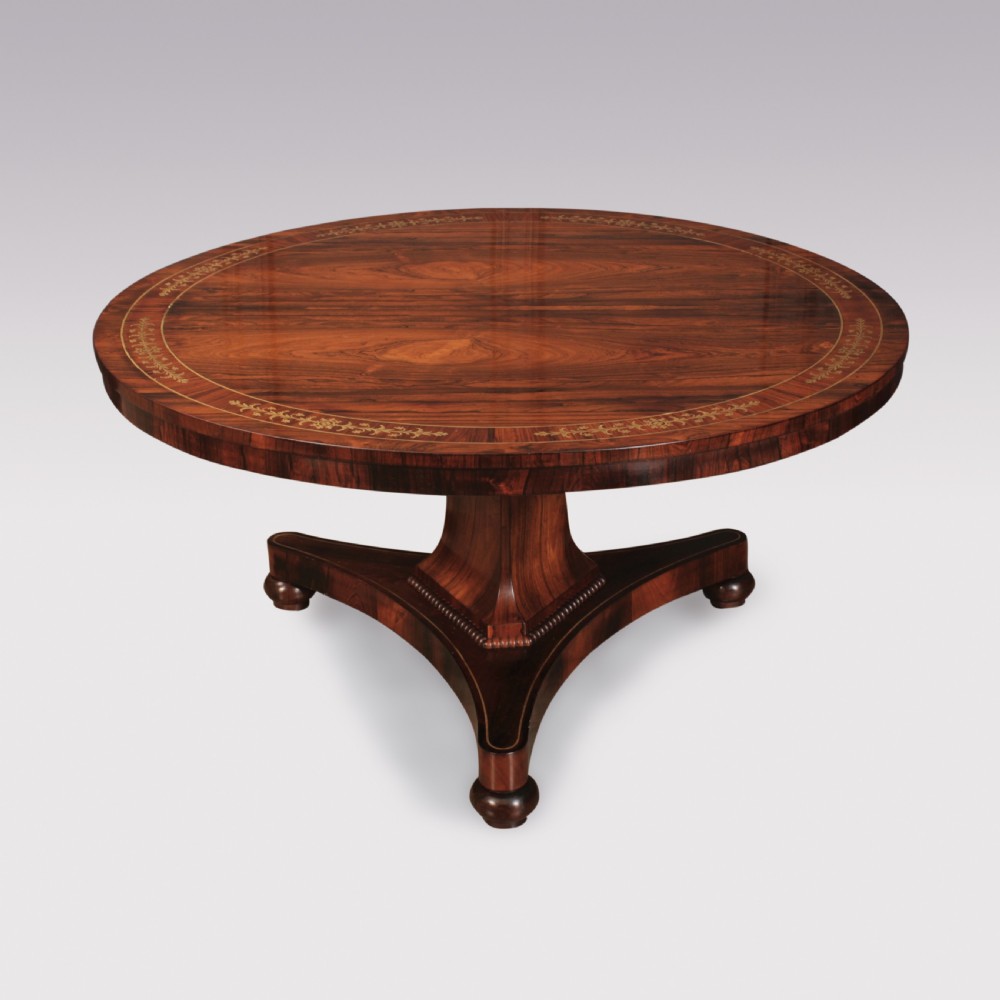A circular Regency rosewood centre table featuring decorative brass inlay around the perimeter and supported by a pedestal on a triform base.
