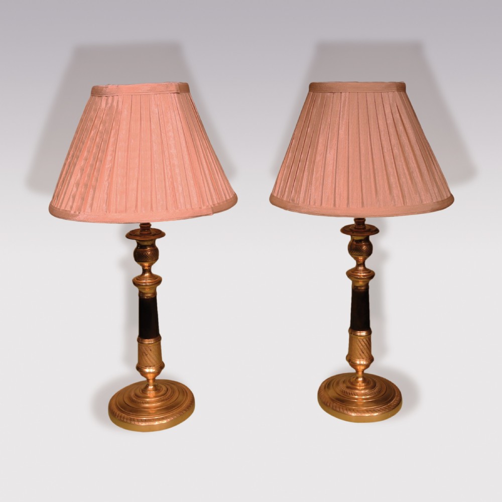 A pair of early 19th-century bronze and ormolu candlestick lamps with dark shafts, gilded bases, and pink pleated fabric lampshades.