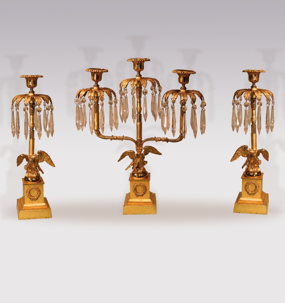 A three-piece Regency period ormolu and gilt bronze candelabra set featuring eagle bases, palm leaf tops, and hanging crystal drops.
