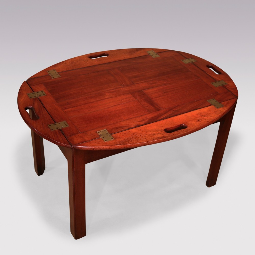 A George III mahogany butlers tray table featuring an oval surface with four folding hinged sides, brass strap hinges, cutout carrying handles, and four square legs.