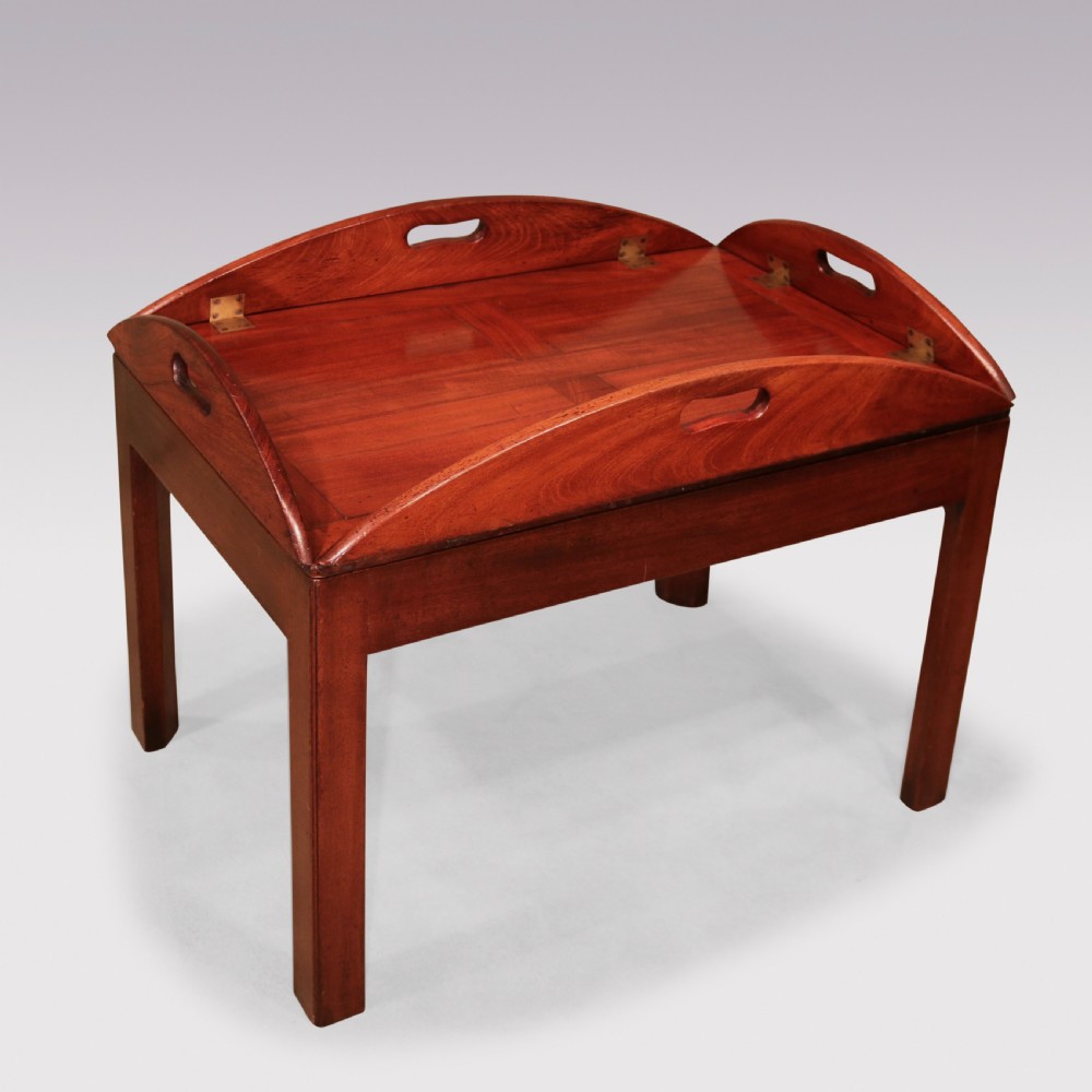 A George III mahogany butlers tray table featuring an oval surface with four folding hinged sides, brass strap hinges, cutout carrying handles, and four square legs. View II