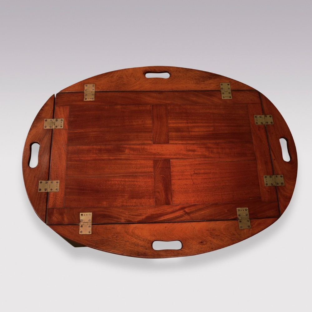 A George III mahogany butlers tray table featuring an oval surface with four folding hinged sides, brass strap hinges, cutout carrying handles, and four square legs. View III