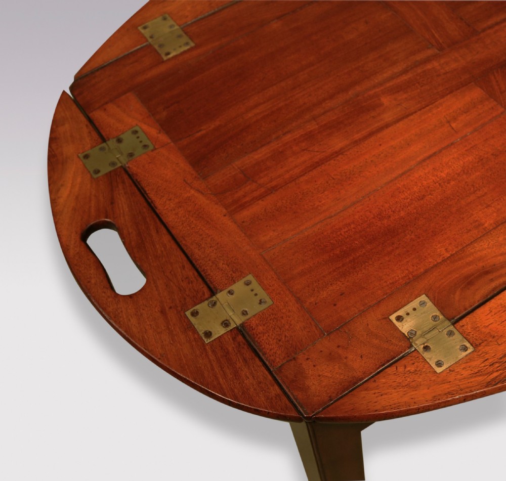 A George III mahogany butlers tray table featuring an oval surface with four folding hinged sides, brass strap hinges, cutout carrying handles, and four square legs. View IV