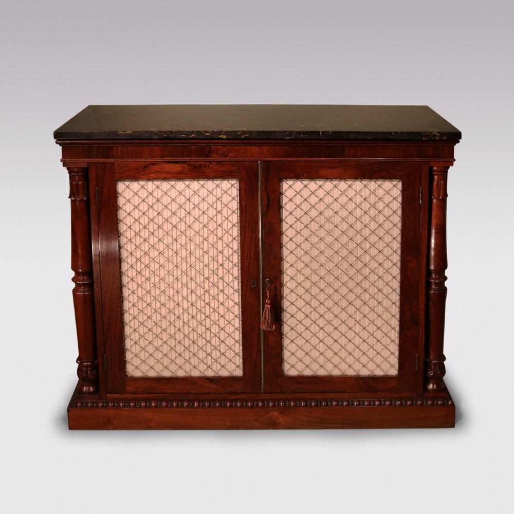 An antique William IV rosewood chiffonier featuring a dark marble top, two brass wire grille doors with pleated fabric backing, and flanking carved side columns.