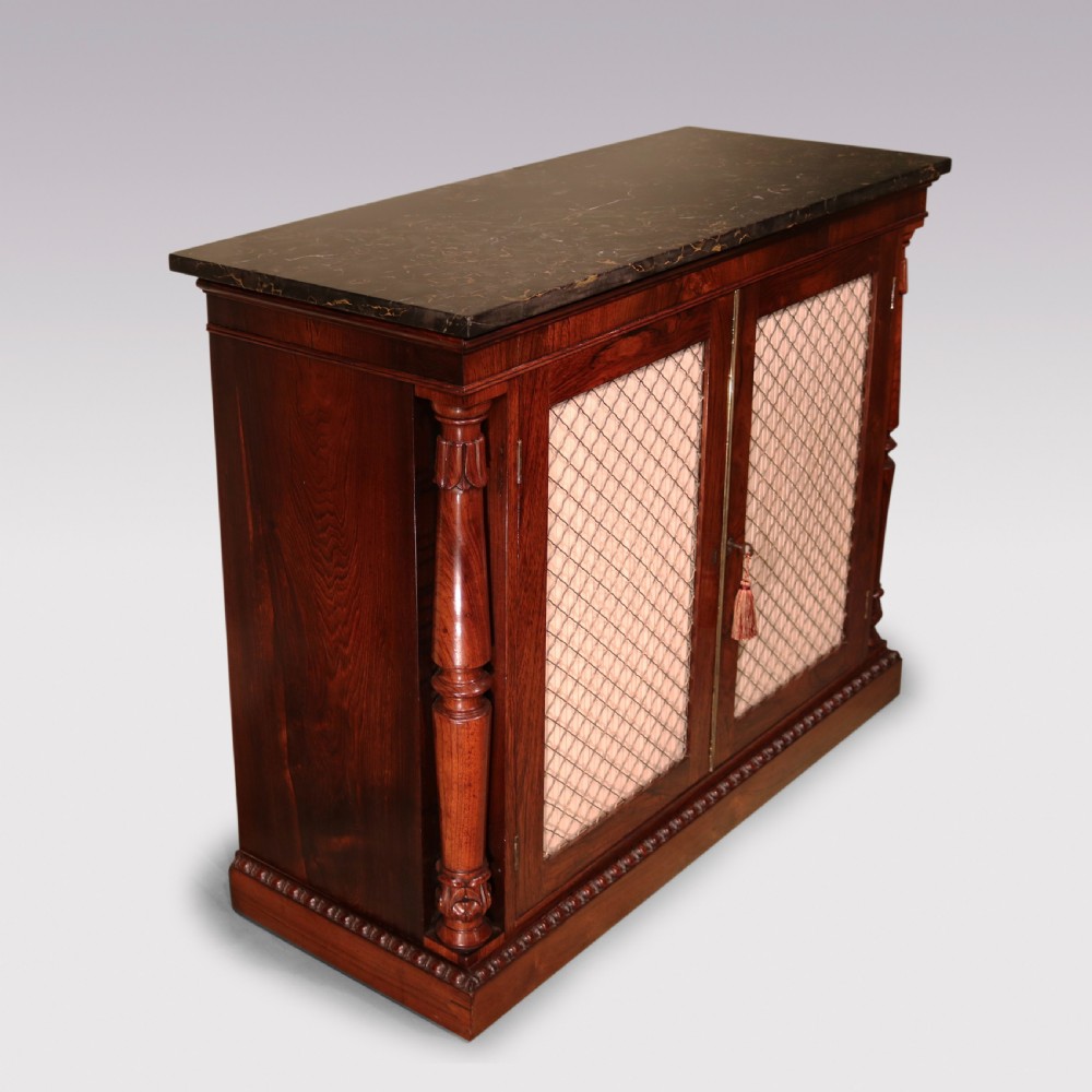 An antique William IV rosewood chiffonier featuring a dark marble top, two brass wire grille doors with pleated fabric backing, and flanking carved side columns. View II