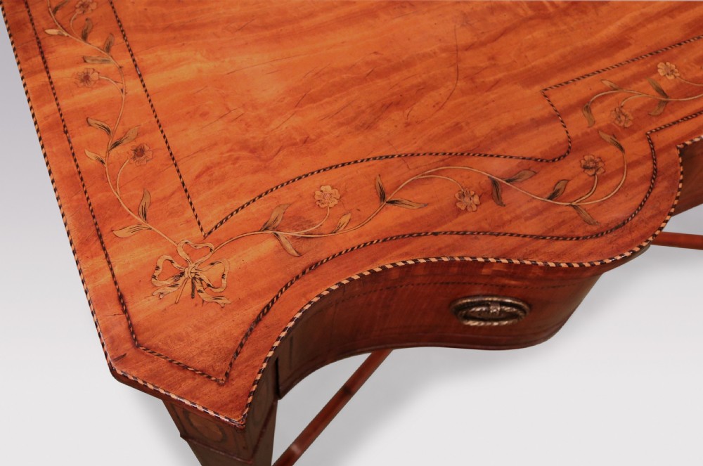 A late 19th-century satinwood hall table featuring a serpentine front, three drawers with brass handles, floral marquetry inlay on square tapered legs, and a central circular under-shelf connected by curved stretchers. View IV