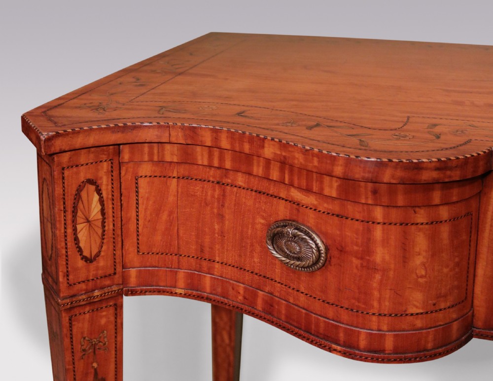 A late 19th-century satinwood hall table featuring a serpentine front, three drawers with brass handles, floral marquetry inlay on square tapered legs, and a central circular under-shelf connected by curved stretchers. View V