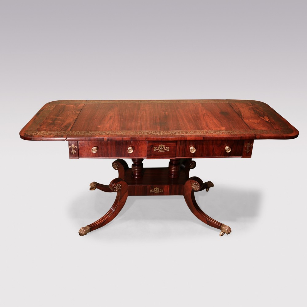 An antique Regency rosewood sofa table with intricate brass inlay, drop-leaf ends, shallow drawers, and swept legs on brass paw castors.