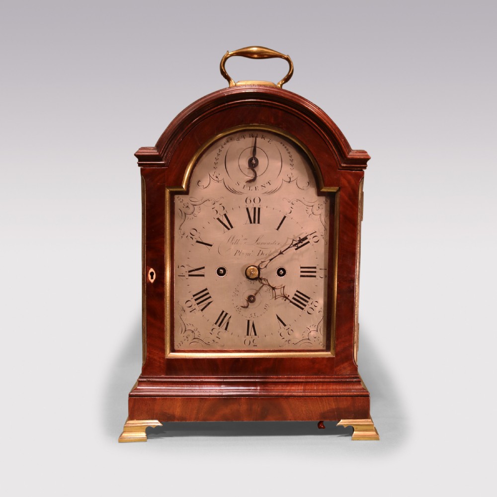 A George III mahogany bracket clock featuring an arched top with a brass carrying handle, an engraved silvered dial with Roman numerals, and brass bracket feet.
