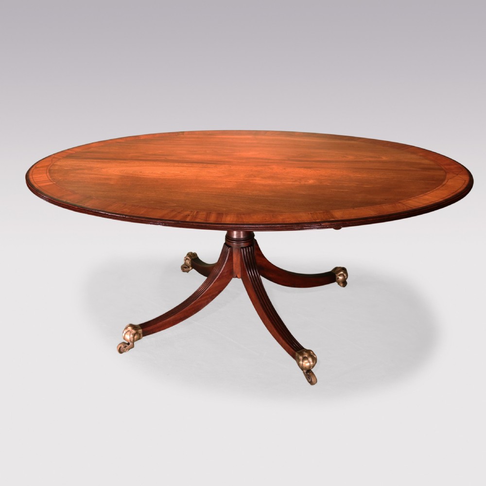 An antique George III oval mahogany breakfast table featuring a figured grain top with crossbanded edging, supported on a central turned pedestal with four splayed legs and brass castors.