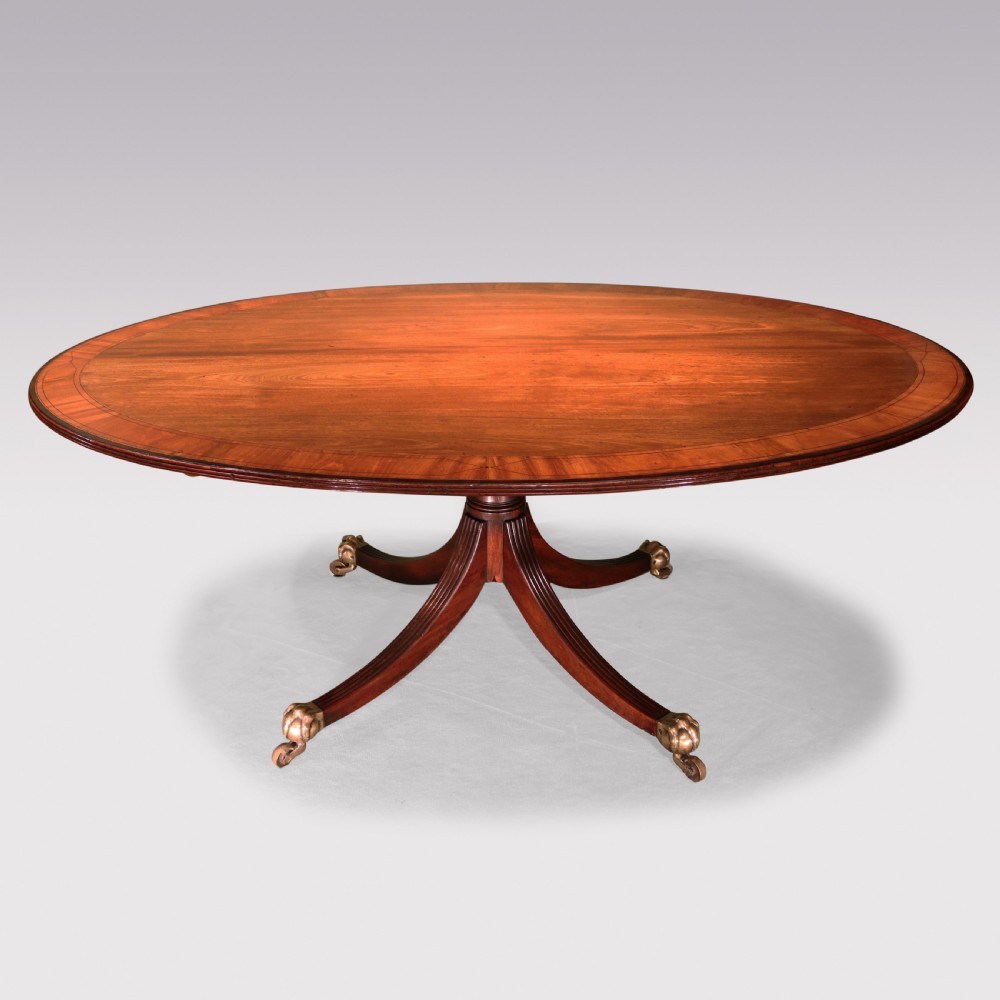 An antique George III oval mahogany breakfast table featuring a figured grain top with crossbanded edging, supported on a central turned pedestal with four splayed legs and brass castors. View II
