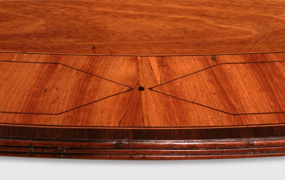 An antique George III oval mahogany breakfast table featuring a figured grain top with crossbanded edging, supported on a central turned pedestal with four splayed legs and brass castors. View III
