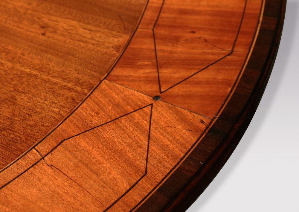 An antique George III oval mahogany breakfast table featuring a figured grain top with crossbanded edging, supported on a central turned pedestal with four splayed legs and brass castors. View IV