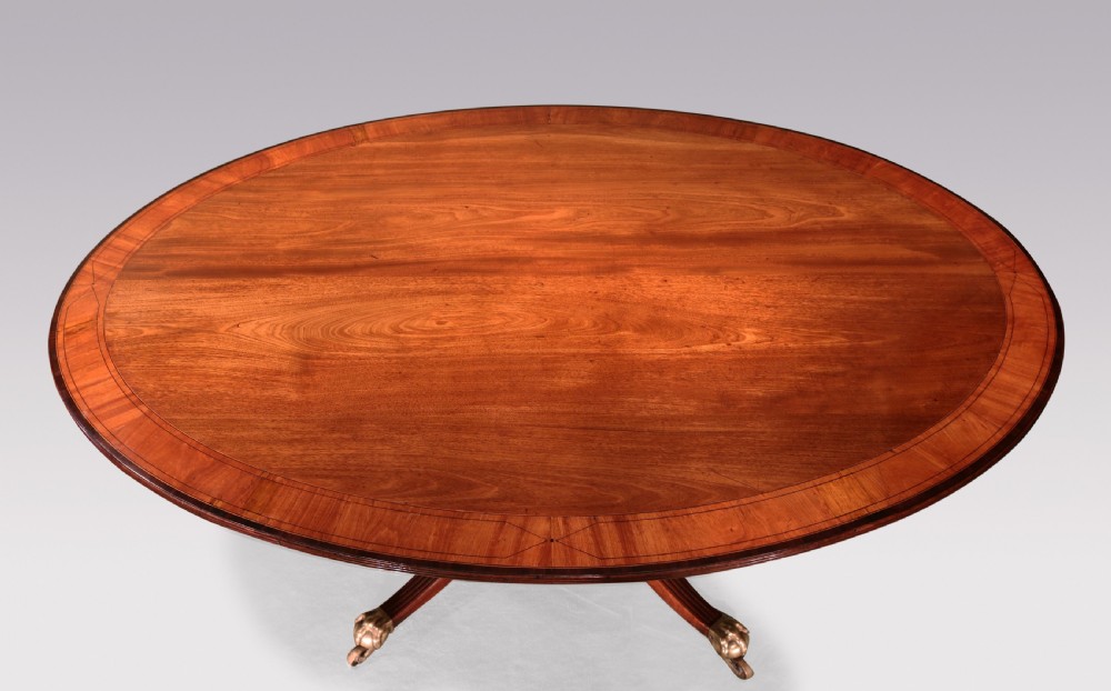 An antique George III oval mahogany breakfast table featuring a figured grain top with crossbanded edging, supported on a central turned pedestal with four splayed legs and brass castors. View V