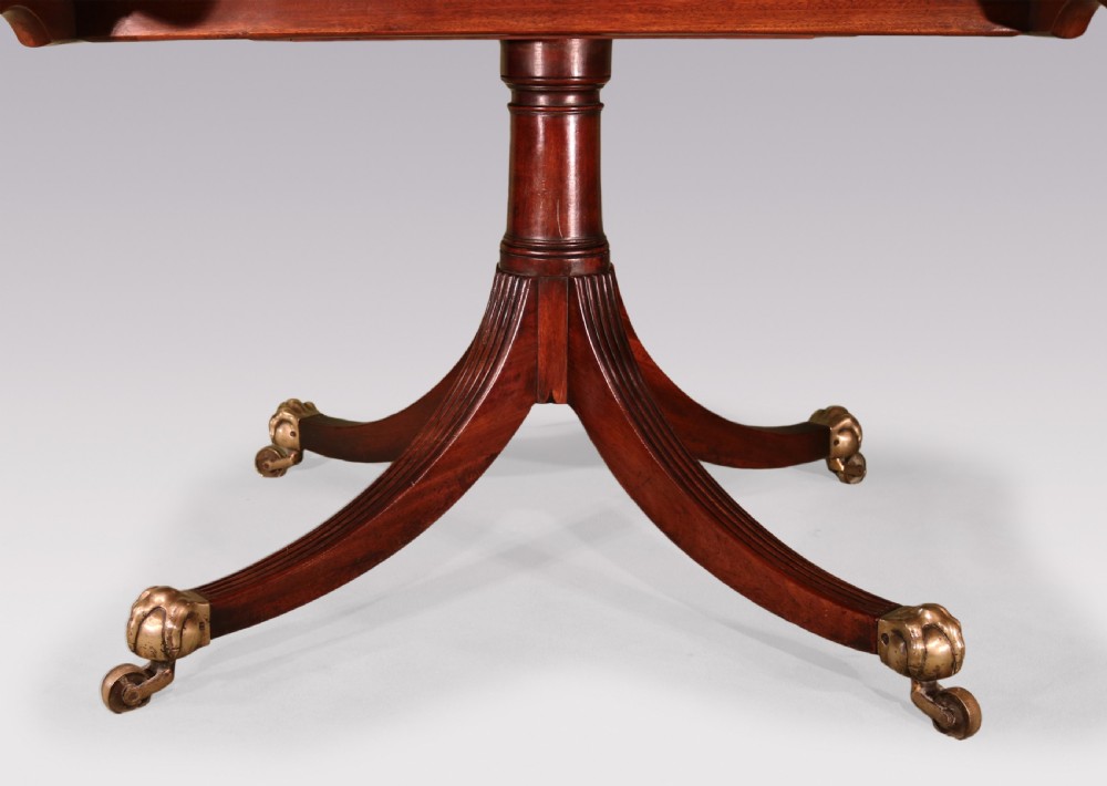 An antique George III oval mahogany breakfast table featuring a figured grain top with crossbanded edging, supported on a central turned pedestal with four splayed legs and brass castors. View VI