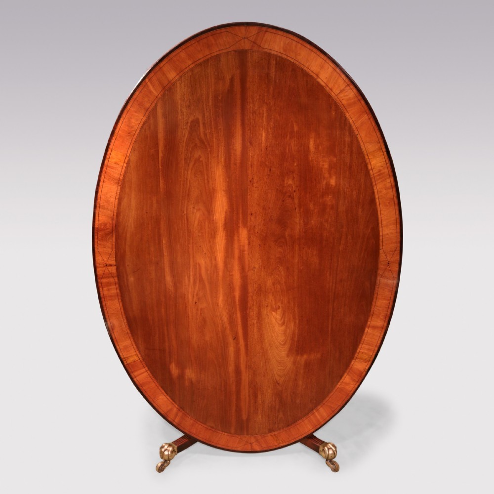 An antique George III oval mahogany breakfast table featuring a figured grain top with crossbanded edging, supported on a central turned pedestal with four splayed legs and brass castors. View VII