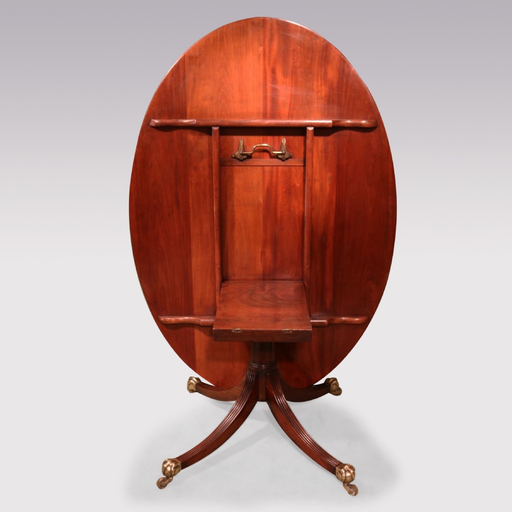 An antique George III oval mahogany breakfast table featuring a figured grain top with crossbanded edging, supported on a central turned pedestal with four splayed legs and brass castors. View VIII