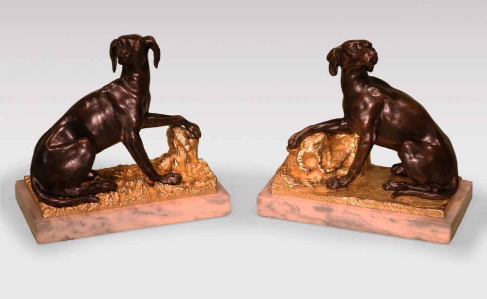 A pair of 19th-century dark bronze sculptures featuring seated hounds resting their paws on gilded ormolu bases depicting pheasants, mounted on rectangular marble plinths.