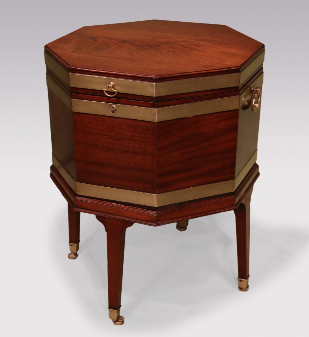 An octagonal George III mahogany wine cooler featuring brass straps, side drop handles, hinged lid, and tapered legs on castors.