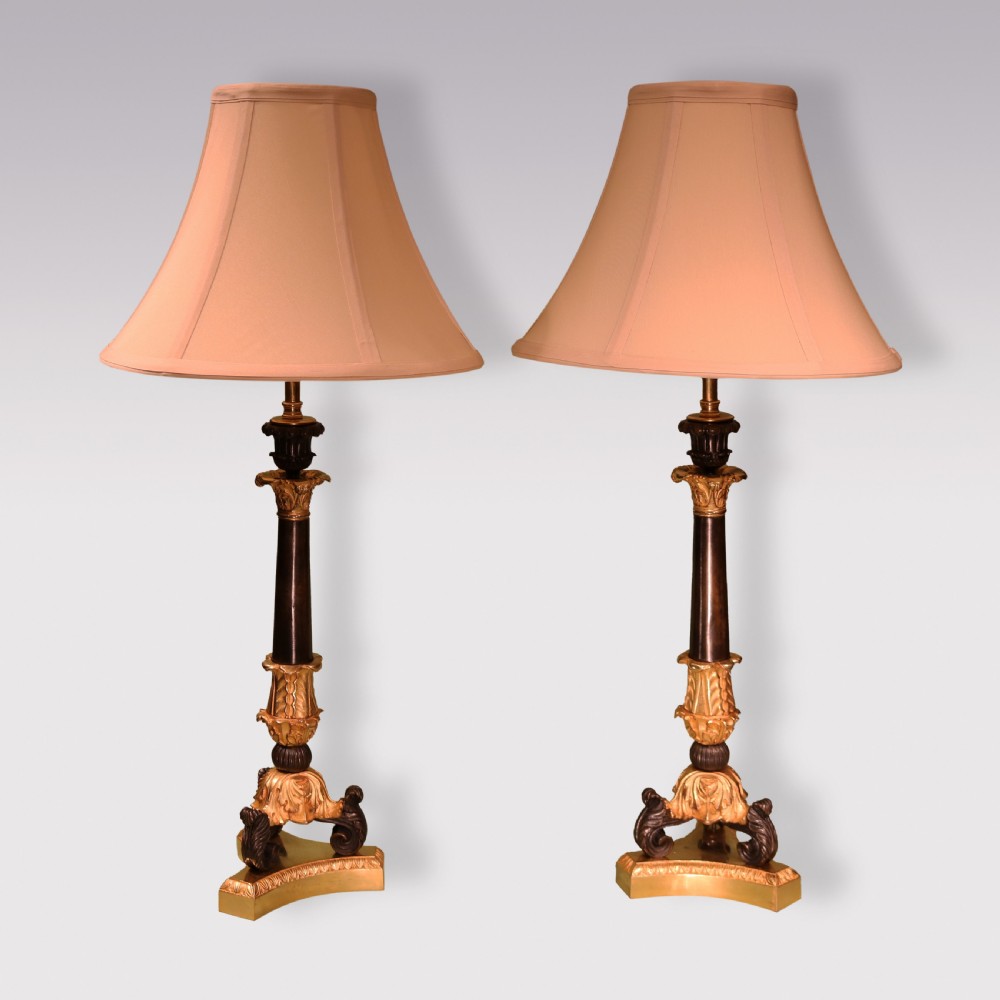 A pair of 19th-century Neoclassical table lamps featuring dark patinated bronze columns, ornate gilt-bronze acanthus leaf mounts, tripod scrolled bases, and flared fabric shades.