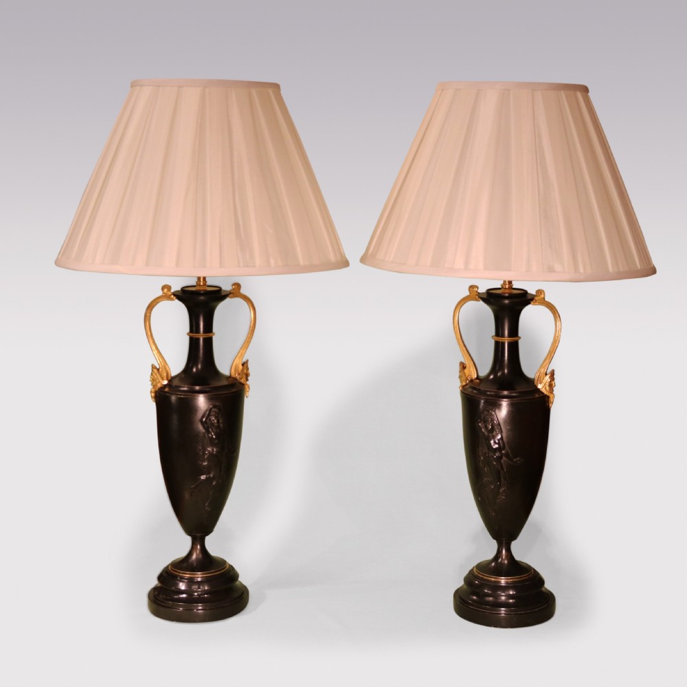 A pair of mid 19th-century dark patinated bronze urn-form table lamps featuring gilt handles, relief figures, circular bases, and cream pleated lampshades.