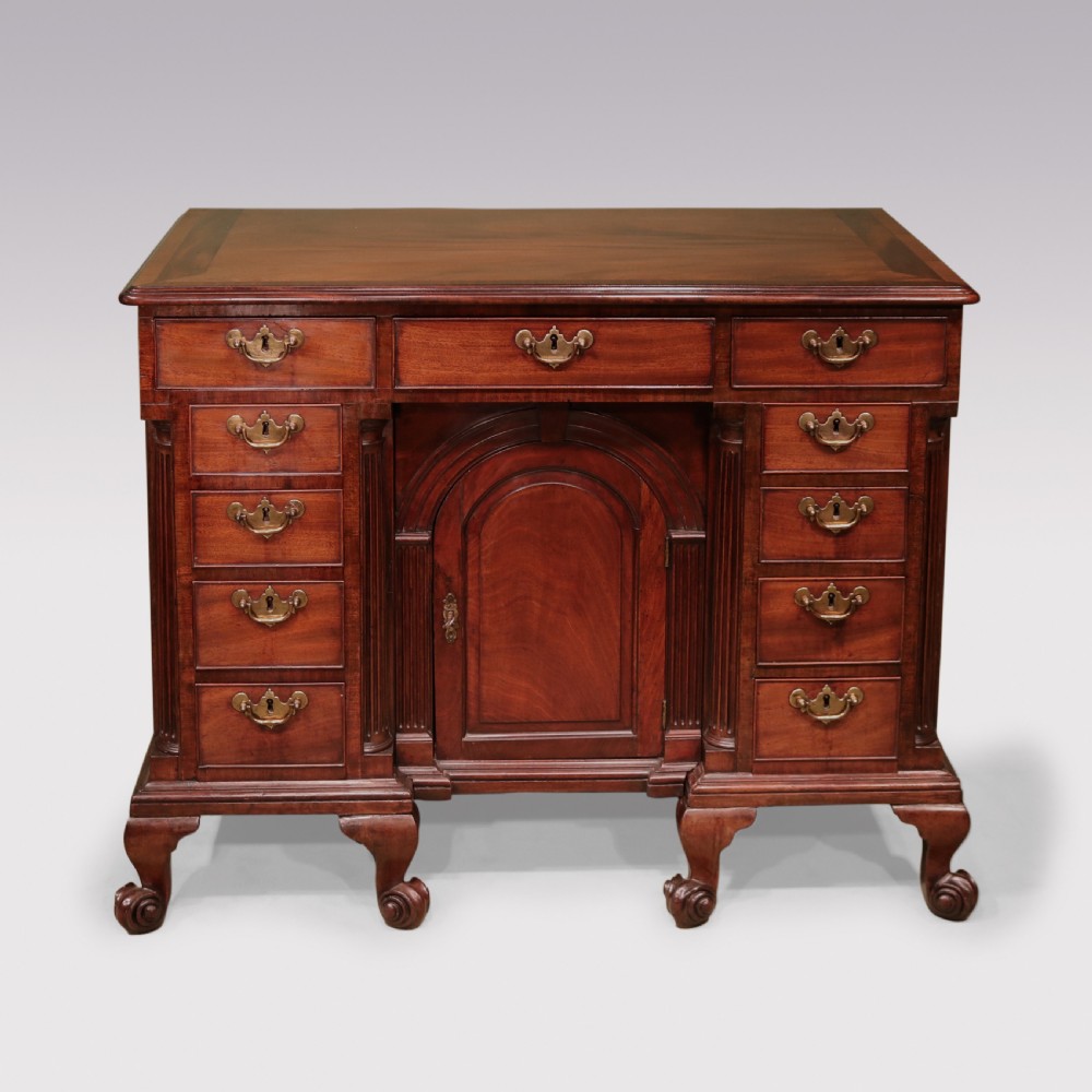 An antique George II mahogany kneehole desk featuring a recessed central cupboard, multiple brass-handled drawers, fluted quarter-columns, and carved scroll feet.