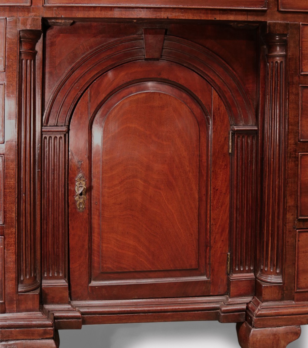 An antique George II mahogany kneehole desk featuring a recessed central cupboard, multiple brass-handled drawers, fluted quarter-columns, and carved scroll feet. View VI
