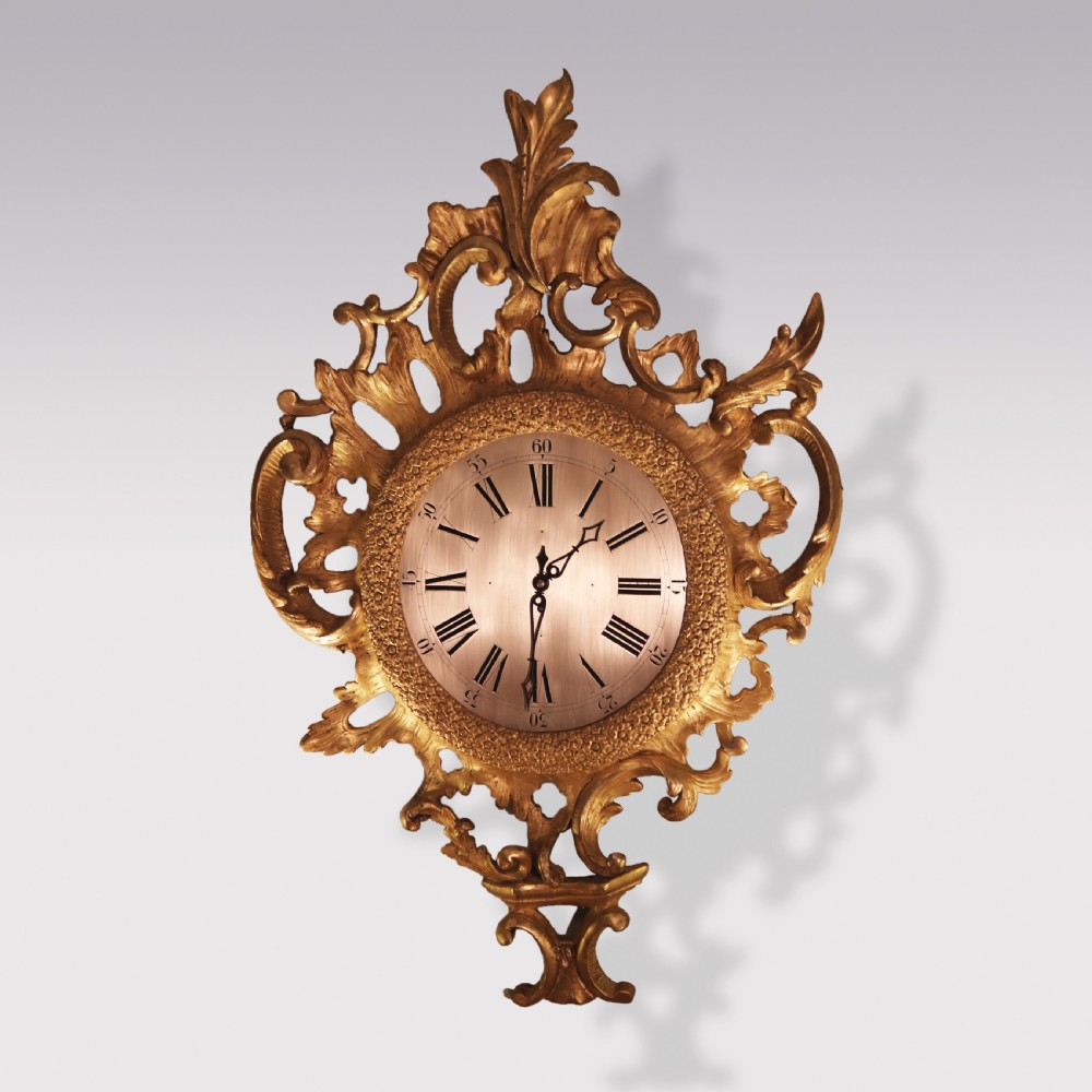 An ornate George III period carved giltwood cartel wall clock featuring asymmetrical rococo scrollwork, acanthus leaf motifs, and a circular dial with Roman numerals.