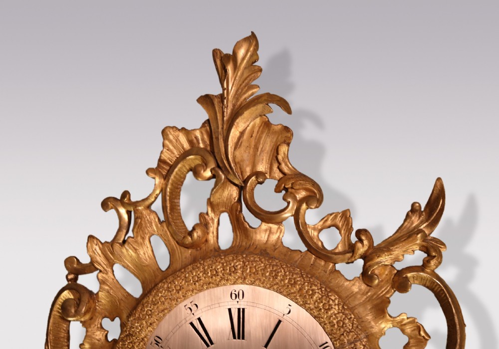 An ornate George III period carved giltwood cartel wall clock featuring asymmetrical rococo scrollwork, acanthus leaf motifs, and a circular dial with Roman numerals. View II