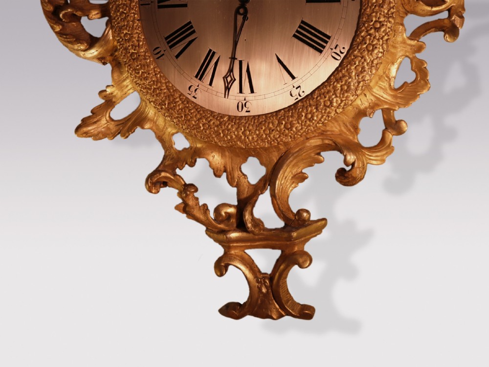An ornate George III period carved giltwood cartel wall clock featuring asymmetrical rococo scrollwork, acanthus leaf motifs, and a circular dial with Roman numerals. View III