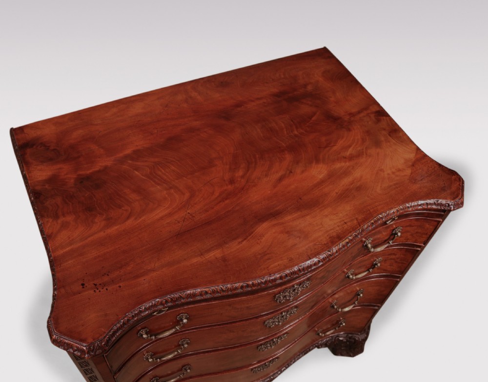 An antique George III mahogany chest of drawers featuring a serpentine front, four graduated drawers with ornamental brass handles, and carved bracket feet. View VI