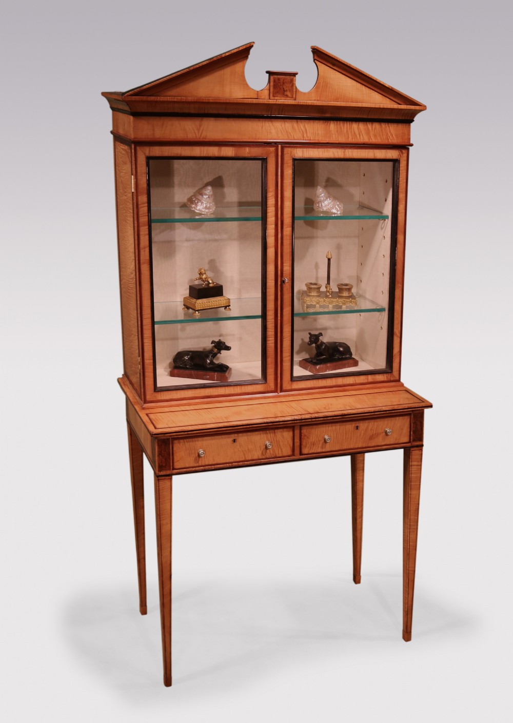 A George III period harewood display cabinet featuring a swan-neck pediment, two glazed doors, two lower drawers, and square tapered legs.
