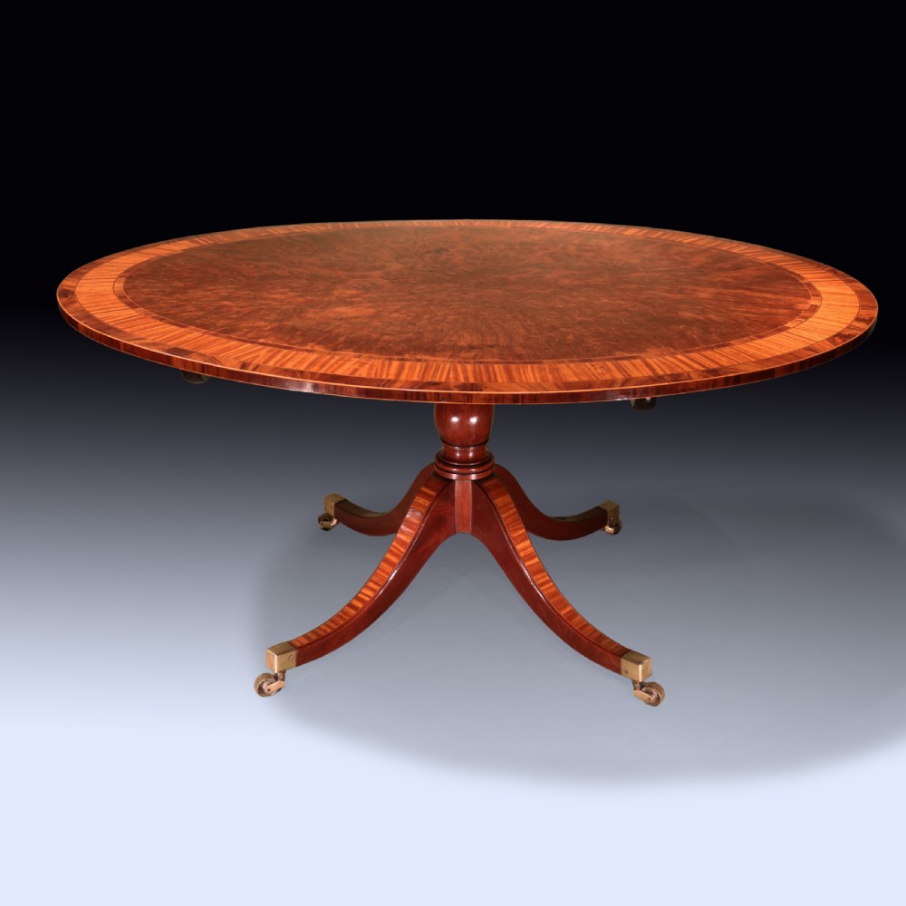 An antique George III oval mahogany breakfast table featuring satinwood crossbanding set on a central pedestal base with four splayed legs and brass castors.