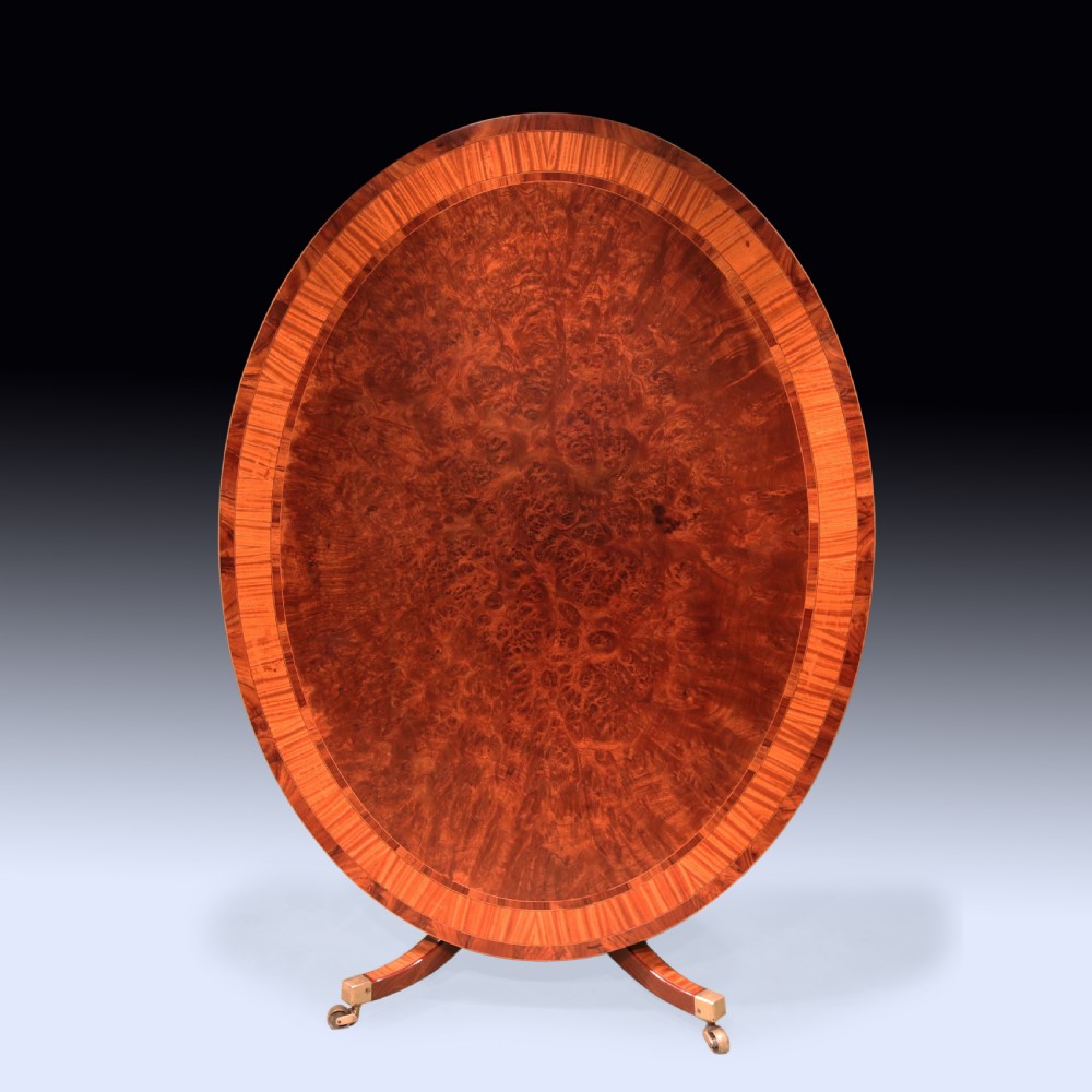 An antique George III oval mahogany breakfast table featuring satinwood crossbanding set on a central pedestal base with four splayed legs and brass castors. View II