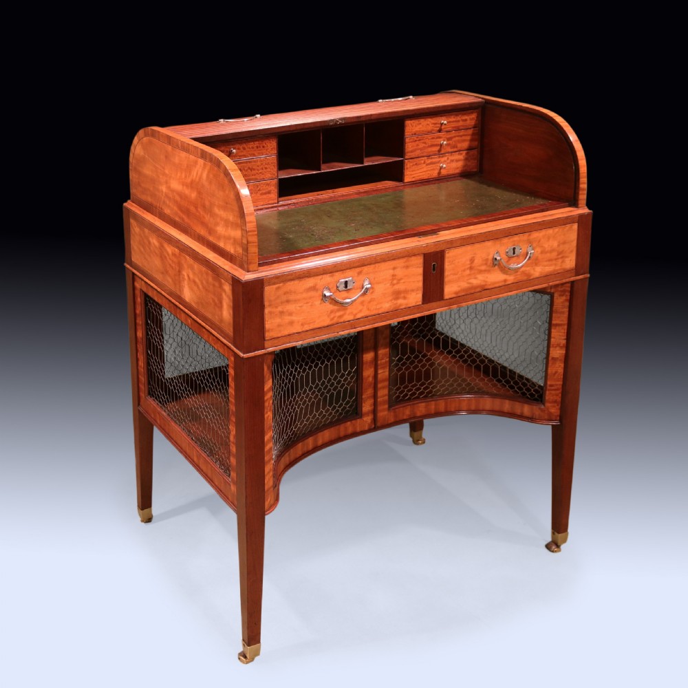 A George III mahogany and satinwood writing table featuring a roll-top compartment, green leather writing surface, inner drawers, and wire mesh lower panels on tapered legs with brass castors.